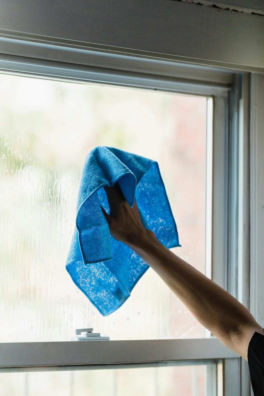 Une main utilisant une serviette bleue pour nettoyer une fenêtre à l'intérieur, mettant en valeur les tâches ménagères.